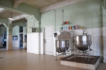 A large industrial kitchen with two massive stainless steel mixing bowls or vats. The room features pale green walls with white tile covering the lower half and a ceiling crisscrossed with exposed beams. Several canned goods and containers are neatly arranged on a shelf above the mixers. The space is well-lit with a lamp hanging from the ceiling.