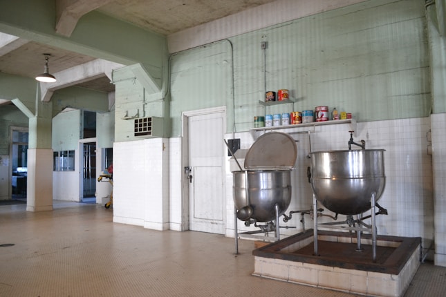 A large industrial kitchen with two massive stainless steel mixing bowls or vats. The room features pale green walls with white tile covering the lower half and a ceiling crisscrossed with exposed beams. Several canned goods and containers are neatly arranged on a shelf above the mixers. The space is well-lit with a lamp hanging from the ceiling.