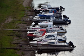 Several motorboats are moored along the calm waters of a river or lake, arranged neatly in a row with grassy areas on the shore. The boats vary in design and color, and some display registration numbers and logos. The atmosphere is relaxed and serene.