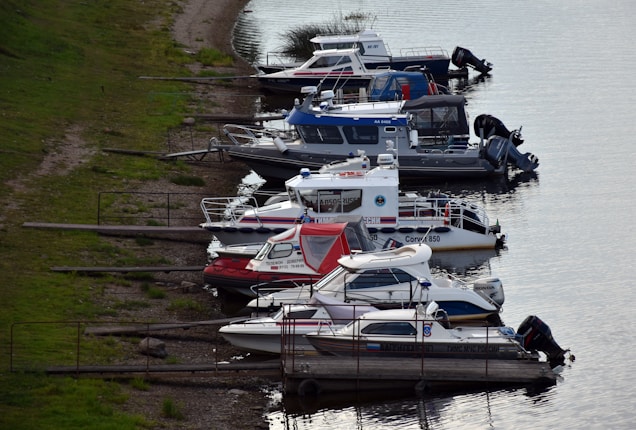 Several motorboats are moored along the calm waters of a river or lake, arranged neatly in a row with grassy areas on the shore. The boats vary in design and color, and some display registration numbers and logos. The atmosphere is relaxed and serene.