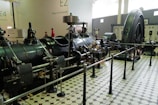 A large industrial machine is on display in a museum setting, featuring dials, gauges, and pipes. The machinery is surrounded by a black rail, and the floor is tiled with a geometric pattern. Walls have numbers and text, while two people, possibly visitors, are seen in the background.