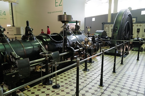 A large industrial machine is on display in a museum setting, featuring dials, gauges, and pipes. The machinery is surrounded by a black rail, and the floor is tiled with a geometric pattern. Walls have numbers and text, while two people, possibly visitors, are seen in the background.