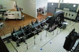 A large industrial engine is displayed in a museum setting, surrounded by a protective barrier. The engine features a massive flywheel and intricate pipework, situated on a vintage tiled floor. Nearby, there is a vintage van and a control panel with meters and gauges along the wall.