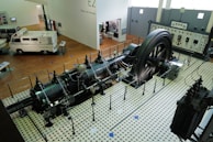 A large industrial engine is displayed in a museum setting, surrounded by a protective barrier. The engine features a massive flywheel and intricate pipework, situated on a vintage tiled floor. Nearby, there is a vintage van and a control panel with meters and gauges along the wall.