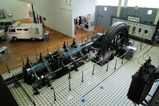 A large industrial engine is displayed in a museum setting, surrounded by a protective barrier. The engine features a massive flywheel and intricate pipework, situated on a vintage tiled floor. Nearby, there is a vintage van and a control panel with meters and gauges along the wall.