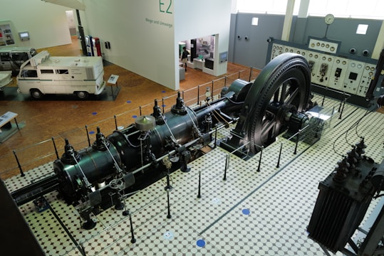 A large industrial engine is displayed in a museum setting, surrounded by a protective barrier. The engine features a massive flywheel and intricate pipework, situated on a vintage tiled floor. Nearby, there is a vintage van and a control panel with meters and gauges along the wall.