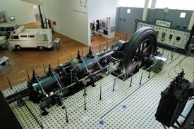 A large industrial engine is displayed in a museum setting, surrounded by a protective barrier. The engine features a massive flywheel and intricate pipework, situated on a vintage tiled floor. Nearby, there is a vintage van and a control panel with meters and gauges along the wall.