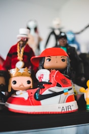 A collection of colorful figurines stands prominently, with the focus on a sneaker-styled toy figure. The shoe features a vivid red and white color scheme with black accents and recognizable branding. Surrounding it are various other figures, each with unique costumes and vibrant details.