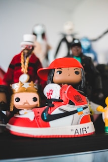 A collection of colorful figurines stands prominently, with the focus on a sneaker-styled toy figure. The shoe features a vivid red and white color scheme with black accents and recognizable branding. Surrounding it are various other figures, each with unique costumes and vibrant details.