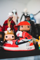 A collection of colorful figurines stands prominently, with the focus on a sneaker-styled toy figure. The shoe features a vivid red and white color scheme with black accents and recognizable branding. Surrounding it are various other figures, each with unique costumes and vibrant details.
