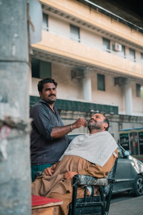 A barber is trimming a customer's beard outdoors, with the customer seated in a reclining chair covered with a towel. They appear to be in a street setting, in front of a building with large windows and visible air conditioning units. The barber is focused on his work, and there is a car parked nearby.