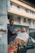 A barber is trimming a customer's beard outdoors, with the customer seated in a reclining chair covered with a towel. They appear to be in a street setting, in front of a building with large windows and visible air conditioning units. The barber is focused on his work, and there is a car parked nearby.