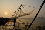 Large fishing nets are suspended on wooden structures along a shoreline at sunset, casting shadows and reflections on the water. The serene scene is complemented by a calm sea and a warm sun hovering near the horizon.
