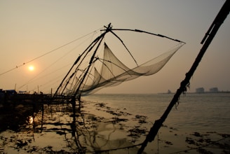 Large fishing nets are suspended on wooden structures along a shoreline at sunset, casting shadows and reflections on the water. The serene scene is complemented by a calm sea and a warm sun hovering near the horizon.