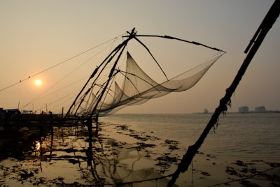Large fishing nets are suspended on wooden structures along a shoreline at sunset, casting shadows and reflections on the water. The serene scene is complemented by a calm sea and a warm sun hovering near the horizon.