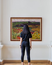 A person stands in a gallery or museum observing a vibrant painting. The artwork is colorful and depicts a landscape with rolling hills and fields, painted in an impressionistic style with bold strokes. The viewer is wearing dark clothing and stands with their back to the camera.