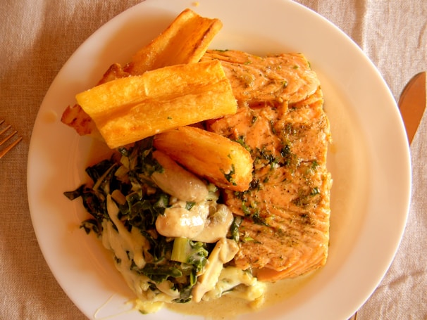 A plate with a serving of grilled salmon, seasoned with herbs, accompanied by deep-fried yuca and a vegetable medley with creamy sauce. The meal is presented on a white plate, placed on a wooden table with a fork and knife set nearby.