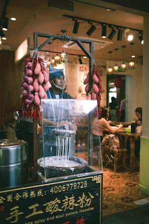 A street food vendor wearing a blue cap operates an apparatus for making noodles, surrounded by strings of pink sausages or similar food items. The scene is set indoors with a warm, ambient lighting and various decorations or lamps hanging from the ceiling. In the background, some patrons are seated at wooden tables, enjoying their meals.
