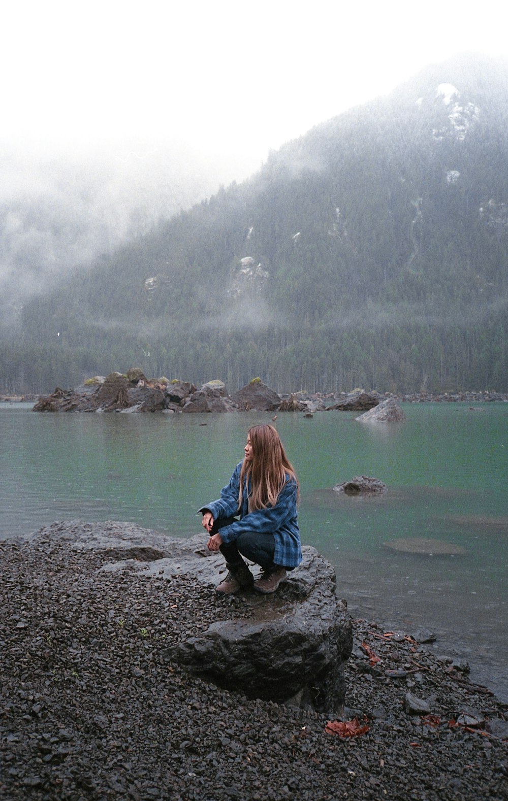 woman in blue jacket sitting on rock by the lake during daytime
