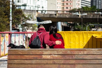 A couple is seated on a wooden bench in an urban environment. Both individuals are wearing matching red hoodies, with one person's arm gently around the other. A black dog is sitting beside them. Behind the couple, a red metal railing and a yellow barrier are visible. In the background, there are buildings and an overpass, indicating a city setting.