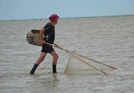 An individual stands waist-deep in water, holding fishing nets attached to long wooden poles. They wear a sun hat, dark clothing, and high waterproof boots. A woven basket is attached to their back, and the overcast sky contributes to a calm, muted atmosphere.