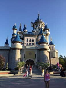 A majestic fairy-tale castle features multiple blue turrets and intricate architectural details. The castle is surrounded by lush green trees and is illuminated by gentle sunlight. People are walking and posing for photos in the foreground, adding a lively atmosphere.