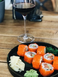 A glass of red wine is placed beside a plate of sushi rolls. The sushi is covered in bright orange roe, and the plate includes garnishes like wasabi, pickled ginger, and a green seaweed salad. Everything is set on a wooden table, presenting a casual dining setting.