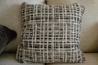 A cushion with a black and white plaid pattern is centered on a beige fabric sofa. The grid-like design on the pillow creates a textured appearance, while another darker pillow is partially visible on the left side of the image. The neutral colors blend harmoniously with the furniture.