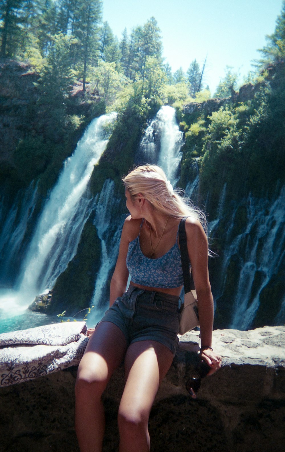 woman in blue tank top and blue denim shorts sitting on rock near waterfalls during daytime