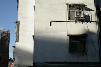 A building facade with a textured, white exterior wall. There are two barred windows with shadows cast from the afternoon sun. An air conditioning unit is partially visible behind one of the windows. The building is simple and utilitarian in design. On the left side, there is part of another structure under construction, wrapped in scaffolding.