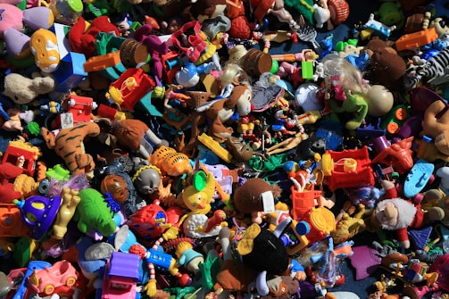 A chaotic assortment of colorful toys piled together, including action figures, plush animals, vehicles, and various shapes and characters. The variety of toys suggests a playful and imaginative setting.