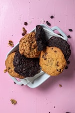 A white ceramic dish contains an assortment of cookies placed on a pink surface. The cookies vary in color and texture, with some appearing to be chocolate chip and others possibly oatmeal or chocolate varieties. Surrounding the dish are scattered cookie crumbs, chocolate chips, and pieces of walnuts.