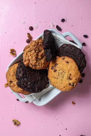 A white ceramic dish contains an assortment of cookies placed on a pink surface. The cookies vary in color and texture, with some appearing to be chocolate chip and others possibly oatmeal or chocolate varieties. Surrounding the dish are scattered cookie crumbs, chocolate chips, and pieces of walnuts.