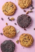 A variety of cookies are arranged on a pink surface. The cookies differ in color and texture, with some being chocolate chip and others appearing to be double chocolate. Scattered around the cookies are ingredients such as chocolate chips, walnuts, coconut flakes, and raisins.