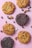 A variety of cookies are arranged on a pink surface. The cookies differ in color and texture, with some being chocolate chip and others appearing to be double chocolate. Scattered around the cookies are ingredients such as chocolate chips, walnuts, coconut flakes, and raisins.