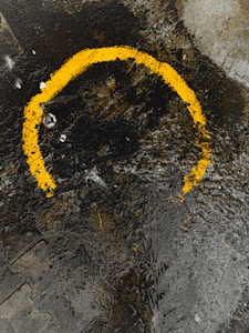 A textured surface of wet asphalt displays a worn, irregular yellow semicircle. The pavement features rough patches, dirt, and small puddles, reflecting a mixture of rugged urban environment and traces of human activity.