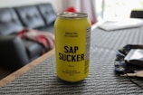 A yellow can of carbonated maple water with the name SAP SUCKER is placed on a textured placemat. The background includes part of a living room with a couch, a blurred red and gray blanket, and soft lighting indicating a casual, indoor setting.