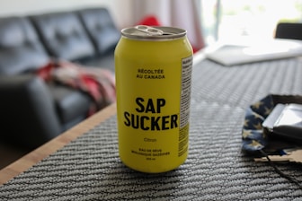 A yellow can of carbonated maple water with the name SAP SUCKER is placed on a textured placemat. The background includes part of a living room with a couch, a blurred red and gray blanket, and soft lighting indicating a casual, indoor setting.