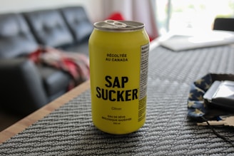 A yellow can of carbonated maple water with the name SAP SUCKER is placed on a textured placemat. The background includes part of a living room with a couch, a blurred red and gray blanket, and soft lighting indicating a casual, indoor setting.