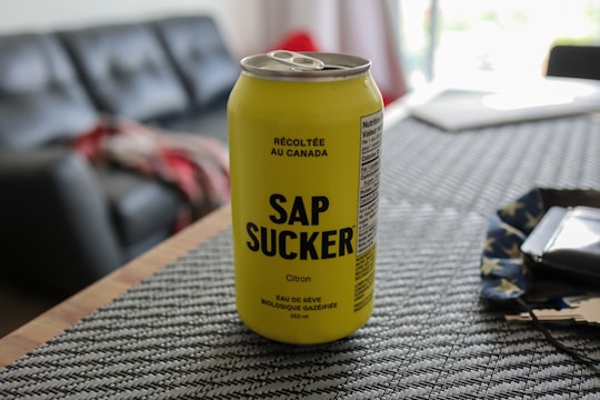 A yellow can of carbonated maple water with the name SAP SUCKER is placed on a textured placemat. The background includes part of a living room with a couch, a blurred red and gray blanket, and soft lighting indicating a casual, indoor setting.