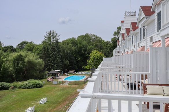 A large white building with multiple balconies overlooking a lush green area with trees and a swimming pool. There are several people lounging by the pool and sitting under umbrellas. The grass is well-maintained, and there are a few white chairs scattered around. The building has white railings and red-striped awnings over the windows.