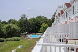 A large white building with multiple balconies overlooking a lush green area with trees and a swimming pool. There are several people lounging by the pool and sitting under umbrellas. The grass is well-maintained, and there are a few white chairs scattered around. The building has white railings and red-striped awnings over the windows.