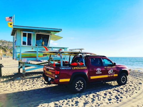 A lifeguard tower and a red lifeguard truck are positioned on a sandy beach. Flags are flying on top of the tower. The ocean is visible on the right, extending to the horizon. The sky is clear and blue.