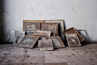 A collection of old, dusty framed photographs and portraits lies haphazardly on a dusty, cracked floor against a worn-out wall. The frames are mostly wooden, and the photographs inside appear faded and aged, depicting indistinct people. The setting looks abandoned and neglected, with dirt and rubble scattered around.