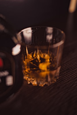 A close-up of a glass filled with a dark amber liquid, likely a type of whiskey, resting on a wooden surface. The edges of the glass are intricately cut, creating a pattern that refracts light. The liquid appears to have ice cubes, and the glass captures a warm, cozy ambiance.