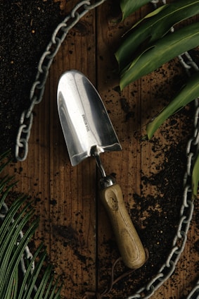 A stainless steel garden trowel with a wooden handle lies on a wooden surface surrounded by dark soil. Green leaves and a metal chain form a border around the scene, suggesting a gardening theme.