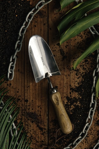 A stainless steel garden trowel with a wooden handle lies on a wooden surface surrounded by dark soil. Green leaves and a metal chain form a border around the scene, suggesting a gardening theme.