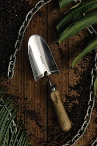 A stainless steel garden trowel with a wooden handle lies on a wooden surface surrounded by dark soil. Green leaves and a metal chain form a border around the scene, suggesting a gardening theme.