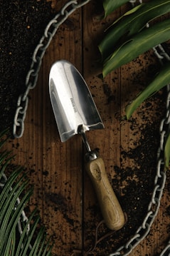 A stainless steel garden trowel with a wooden handle lies on a wooden surface surrounded by dark soil. Green leaves and a metal chain form a border around the scene, suggesting a gardening theme.