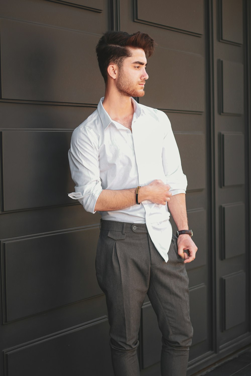 白いドレスシャツと灰色のドレスパンツを着た男が黒い木製のドアのそばに立っている